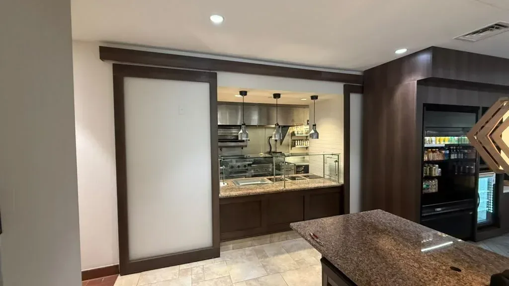 Dark wood buffet cabinetry with a frosted sliding panel framing a commercial kitchen pass-through, with stone countertops and pendant lighting visible